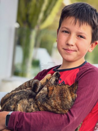 Foto de Primer plano retrato de amoroso adolescente caucásico chico sonriendo y llevando gato mientras sentado en patio - Imagen libre de derechos