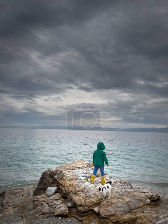 Foto de Vista trasera del niño con abrigo y botas de goma mirando al mar mientras está de pie con el gato en la roca contra las nubes de tormenta - Imagen libre de derechos