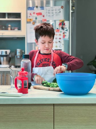 Foto de Niño caucásico usando delantal picando brócoli en la tabla de cortar mientras se aprende a cocinar en la cocina en casa - Imagen libre de derechos