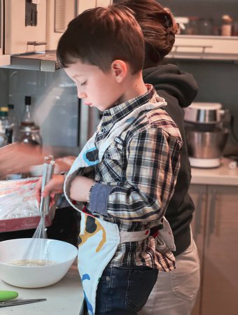 Foto de Vista lateral del lindo chico caucásico usando delantal mezclando masa en un tazón mientras hornea con la madre en la cocina - Imagen libre de derechos