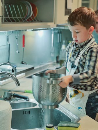 Foto de Lindo chico caucásico drenando agua del colador sobre el fregadero mientras aprende a cocinar en la cocina en casa - Imagen libre de derechos