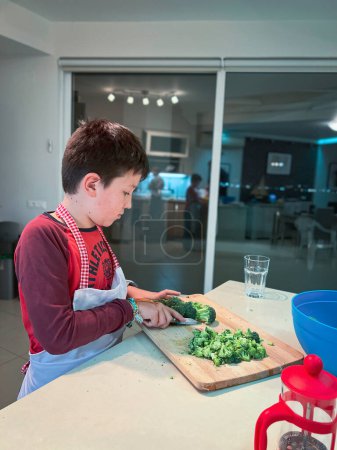 Foto de Vista lateral del adolescente caucásico usando delantal picando brócoli en la tabla de cortar en la cocina en casa - Imagen libre de derechos