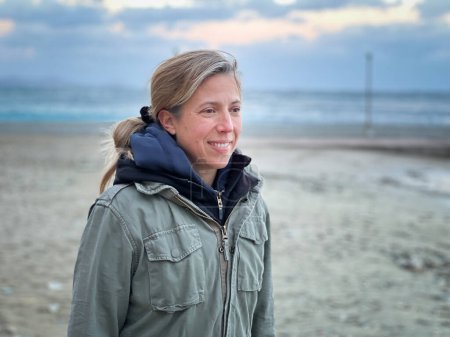 Foto de Sonriente mujer blanca mediana adulta usando abrigo de invierno mirando hacia otro lado mientras está de pie en la playa contra el cielo nublado - Imagen libre de derechos