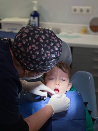 Foto de Odontóloga con gorra floral y guantes quirúrgicos examinando dientes de niño sentado en silla en clínica dental - Imagen libre de derechos