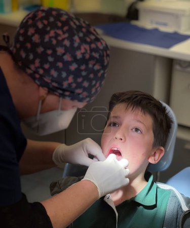 Foto de Dentista femenina con gorra floral examinando los dientes de un adolescente sentado en una silla en una clínica dental - Imagen libre de derechos