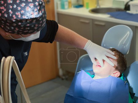 Foto de Dentista femenina con gorra floral sosteniendo la boca del niño caucásico con la mano mientras examina en el hospital dental - Imagen libre de derechos