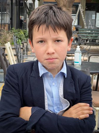 Foto de Retrato de primer plano de un chico caucásico serio usando traje con los brazos cruzados sentado en el restaurante de la acera - Imagen libre de derechos