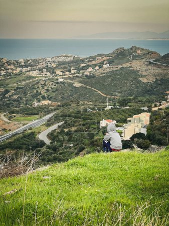 Foto de Vista trasera del niño caucásico con chaqueta encapuchada mirando a la vista mientras está sentado en la montaña cubierta de hierba contra el cielo - Imagen libre de derechos