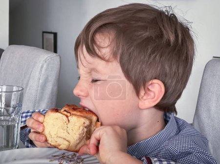 Foto de Vista lateral del chico caucásico con los ojos cerrados tomando un bocado de pan mientras desayuna en la mesa de comedor - Imagen libre de derechos