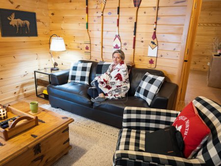 Foto de Retrato de mujer adulta mediana caucásica envuelta en manta relajante en sofá en cabaña de madera mientras disfruta de vacaciones durante el invierno - Imagen libre de derechos