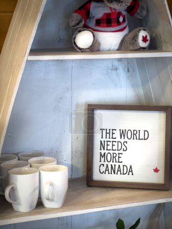 Foto de Primer plano del marco con el mundo necesita más texto de Canadá por tazas blancas dispuestas en estante de madera en casa - Imagen libre de derechos