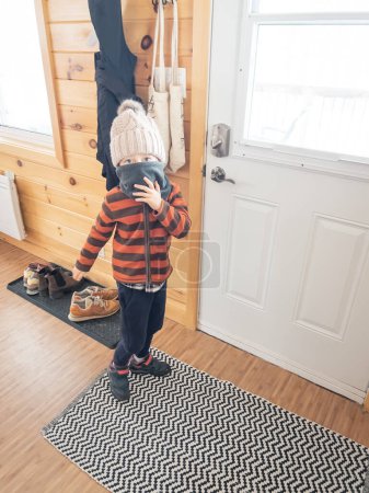 Foto de Retrato de niño caucásico juguetón escondiendo la cara con bufanda mientras está de pie junto a la puerta cerrada en casa - Imagen libre de derechos
