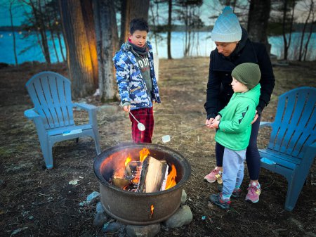 Photo for Caucasian mother with sons roasting marshmallows over campfire while standing in woodland during sunset - Royalty Free Image