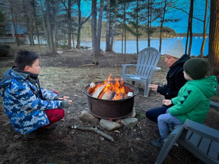 Photo for Caucasian mother with sons roasting marshmallows over campfire while sitting on chairs in forest during sunset - Royalty Free Image