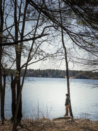 Foto de Vista lateral del niño mirando a la vista mientras está de pie junto al tronco del árbol a orillas del lago contra el cielo en el bosque durante el día soleado - Imagen libre de derechos