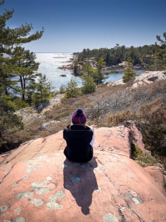Foto de Vista trasera de la mujer adulta que lleva ropa de abrigo mirando al mar mientras está sentada en la formación de rocas durante el día soleado - Imagen libre de derechos