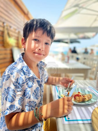 Foto de Retrato de niño caucásico sonriente lindo comiendo rebanada de pizza en la mesa mientras está sentado en el restaurante al aire libre durante el fin de semana - Imagen libre de derechos