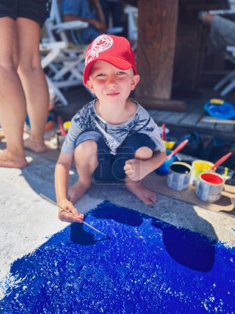 Foto de Retrato de lindo niño caucásico con gorra y pintura de color azul con pincel en la pasarela durante el día soleado - Imagen libre de derechos