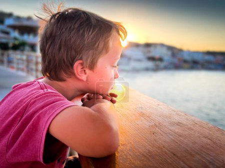 Foto de Vista lateral del niño lindo reflexivo mirando al mar mientras descansa la cabeza sobre una barandilla de madera contra el cielo durante el atardecer - Imagen libre de derechos