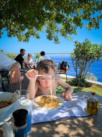 Foto de Retrato de un chico caucásico lindo sin camisa comiendo espaguetis con tenedor en la mesa mientras disfruta del fin de semana en un restaurante al aire libre - Imagen libre de derechos