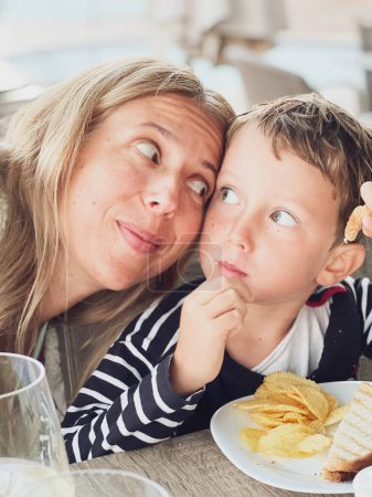 Foto de Primer plano de la mujer sonriente está mirando hijo comiendo chico lindo comer sándwich y patatas fritas en la mesa en el restaurante - Imagen libre de derechos