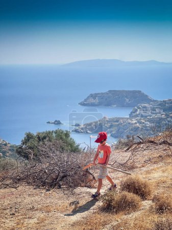 Foto de Niño caucásico con gorra y pala mientras camina en la montaña contra el mar y el cielo durante el día soleado - Imagen libre de derechos