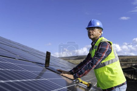 Téléchargez les photos : Portrait de l'ingénieur gestionnaire masculin dans la vérification du casque de sécurité avec ordinateur un fonctionnement du système de panneaux solaires à la station solaire - en image libre de droit