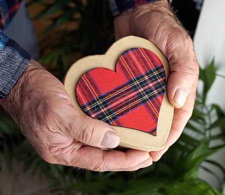 Foto de Gift box in shape of heart of red tartan pattern in hands of old man photo illustration - Imagen libre de derechos