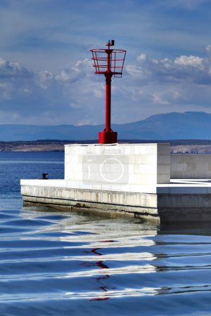 Roter metallischer Leuchtturm auf weißem Steinpier mit Reflexion auf der welligen Meeresoberfläche