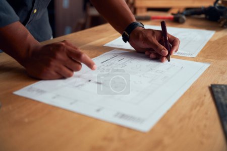 Photo for Close-up of multiracial young man writing with pencil and pointing at paper in woodworking factory - Royalty Free Image