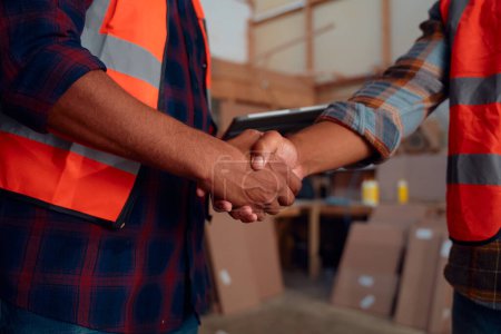 Photo for Close up of multiracial men wearing reflective clothing while shaking hands at woodworking factory - Royalty Free Image