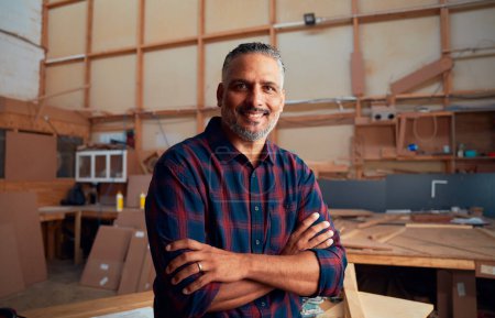 Porträt eines glücklichen, multirassischen erwachsenen Mannes in kariertem Hemd mit verschränkten Armen in einer holzverarbeitenden Fabrik