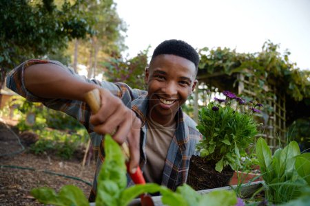 Foto de Feliz joven negro mirando a la cámara y sonriendo mientras usa la pala en el parterre de flores en el vivero de plantas - Imagen libre de derechos