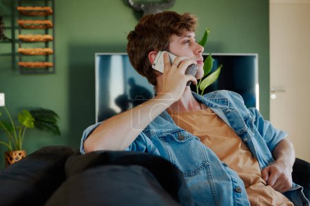 Téléchargez les photos : Vue du profil du jeune homme caucasien en vêtements décontractés avec téléphone portable à l'oreille sur canapé dans le salon à la maison - en image libre de droit