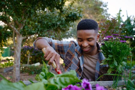 Téléchargez les photos : Heureux jeune homme noir portant chemise à carreaux souriant tout en utilisant bêche dans le lit de fleurs dans le centre de jardin - en image libre de droit