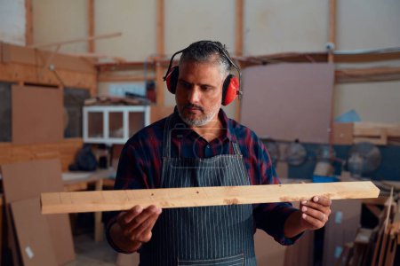 Foto de Multiracial hombre adulto medio usando orejeras mientras sostiene pieza de madera en fábrica de carpintería - Imagen libre de derechos