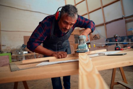 Foto de Multiracial hombre adulto medio usando orejeras usando herramienta eléctrica en tablón de madera en fábrica de carpintería - Imagen libre de derechos