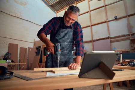 Foto de Multiracial hombre adulto medio sonriendo mientras se utiliza el taladro en la madera junto a la tableta digital en la fábrica de carpintería - Imagen libre de derechos