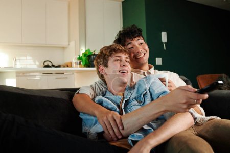 Foto de Feliz joven pareja gay riendo mientras mira hacia otro lado y sosteniendo control remoto en el sofá en la sala de estar en casa - Imagen libre de derechos
