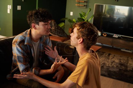 Foto de Joven pareja gay vistiendo ropa casual cara a cara mientras discute y hace gestos en el sofá en casa - Imagen libre de derechos
