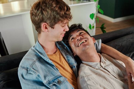Photo for Happy young gay couple smiling while relaxing face to face on sofa in living room at home - Royalty Free Image