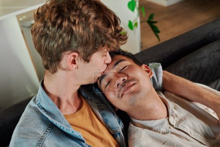 joven gay pareja en casual ropa besos con ojos cerrado en sofá en sala de estar en casa