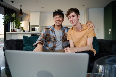 Photo for Young gay couple sitting on sofa and smiling during video call on laptop in living room at home - Royalty Free Image