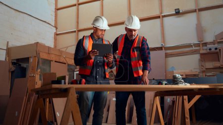 Téléchargez les photos : Hommes multiraciaux dans le casque de sécurité regardant tablette numérique à côté des outils électriques dans l'usine de travail du bois - en image libre de droit