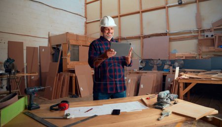 Foto de Multiracial hombre adulto medio en casco de seguridad sonriendo mientras se utiliza tableta digital por mesa en la fábrica de carpintería - Imagen libre de derechos
