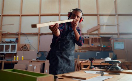 Foto de Multiracial hombre adulto medio usando orejeras examinando madera al lado de herramientas eléctricas en la fábrica de carpintería - Imagen libre de derechos