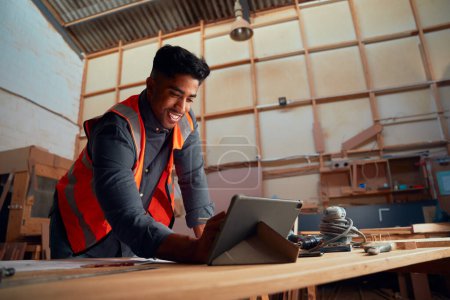 Téléchargez les photos : Jeune homme multiracial en écriture réfléchie de vêtements avec stylo numérisé sur tablette numérique dans l'usine de travail du bois - en image libre de droit