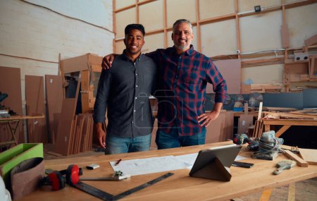 Photo for Group portrait of two multiracial men smiling with arm around behind table in woodworking factory - Royalty Free Image