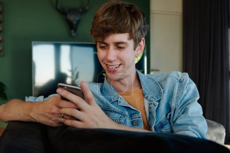 Junger kaukasischer Mann in lässiger Kleidung lächelt, während er im heimischen Wohnzimmer mit dem Handy SMS schreibt