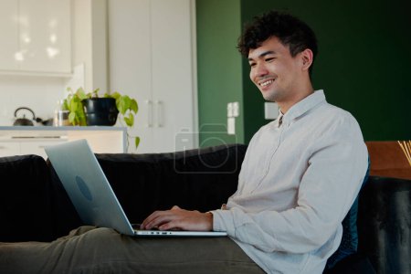 Photo for Happy young multiracial man in shirt using laptop while sitting with feet up on sofa in living room at home - Royalty Free Image